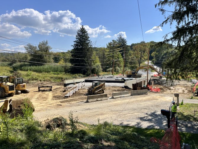 road construction progress photo. A roadway intersection has been removed to show work progressing below the road surface. Multiple pieces of construction equipment are stationed around the work area. The sky is blue with a large puffy cloud in the upper left. The trees are still green in late September.